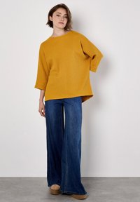 Pull oversized en maille texturée jaune moutarde avec des manches 3/4, associé à un jean en denim bleu à jambes larges et des chaussures à plateforme marron clair.