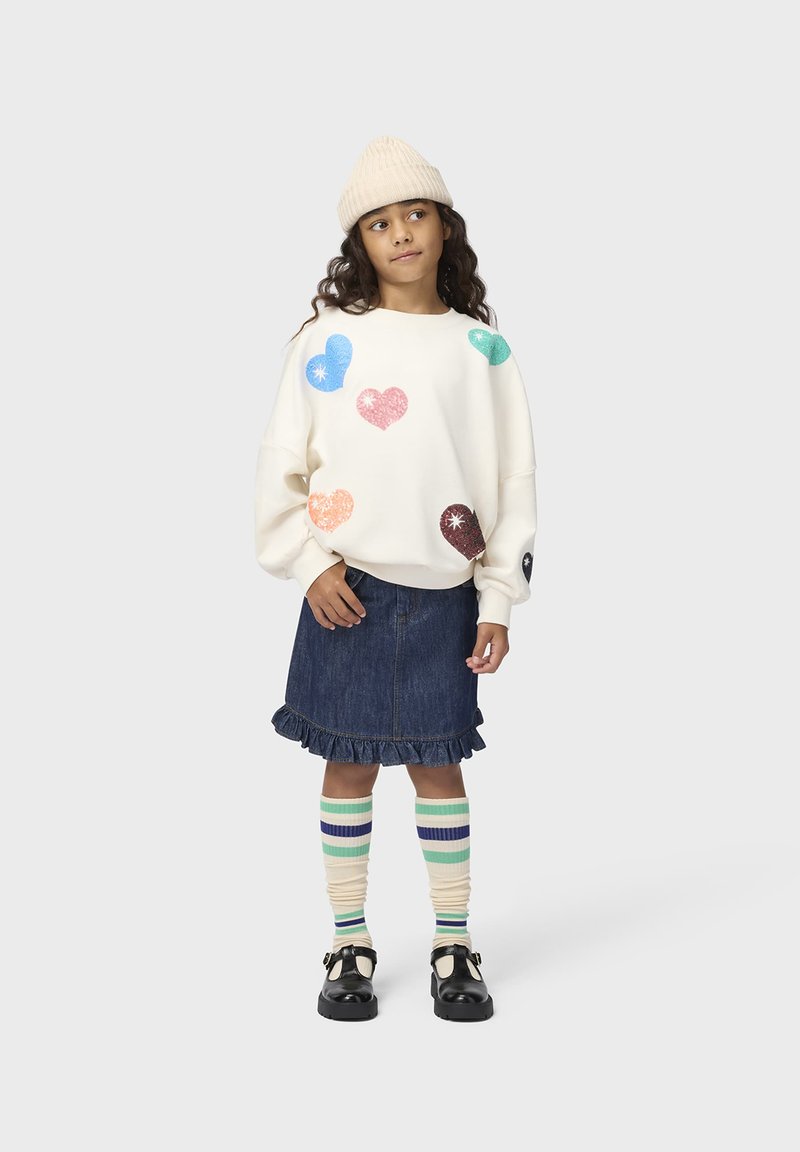 Fille portant un pull blanc avec des cœurs colorés, une jupe en jean avec un ourlet à volants, des chaussettes rayées montant jusqu'aux genoux, des chaussures noires et un bonnet en tricot beige.