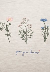 Camisa gris claro con flores bordadas en rosa y azul. Texto abajo: "haz crecer tus sueños" en azul, con un pequeño acento en forma de estrella rosa.