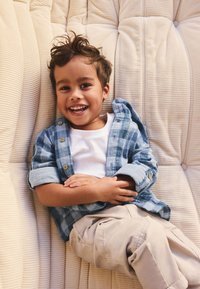 Jeune garçon souriant aux cheveux bouclés portant une chemise à carreaux bleue et un pantalon beige, allongé détendu sur une surface beige rembourrée.