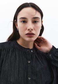 Tmavě modrá džínsová košile s kulatým límcem, zapínáním na knoflíky a nařasenými detaily na ramenou. Texturovaná tkanina s jemným leskem.