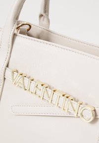 Beige läderhandväska med en framträdande logotyp i guldton "VALENTINO", slät textur, dubbla handtag och minimalistisk design.