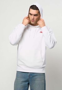 Hombre con sudadera blanca con capucha y un logo rojo, tirando de la capucha sobre su cabeza, de pie contra un fondo claro y liso.