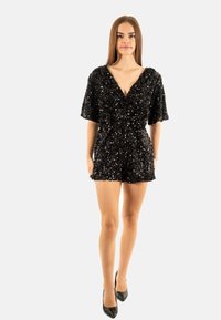 Mono corto negro con lentejuelas, escote en V profundo, mangas cortas y cintura ajustada. La textura es brillante y lustrosa, combinado con tacones negros.