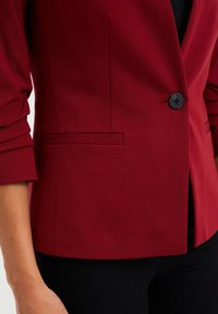 Blazer entallado rojo hecho de tejido suave, con un solo botón negro y puños estructurados, detalle de bolsillo lateral y una silueta elegante.