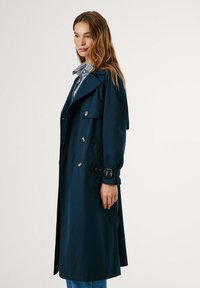 Manteau trench bleu marine en tissu lisse, avec une taille ceinturée, un grand col, des accents de boutons et des poches latérales.