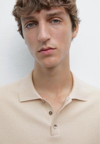 Beige ribbet polo-shirt med krave og to knapper, lavet af blødt stof. Modellen har kort, rodet hår mod en lys baggrund.