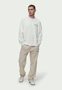 Camisa branca de manga longa com um logótipo preto, combinada com calças de sweat de tom bege claro. O conjunto apresenta um corte descontraído e texturas suaves.