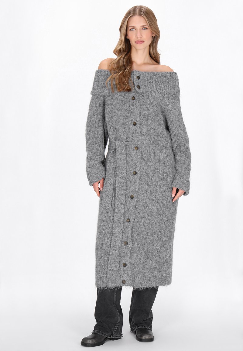 Rochie tricotată gri, cu umerii descoperiți, cu mâneci lungi, nasturi pe partea din față și un cordon la talie. Textura este moale și pufoasă, cu o margine franjurată.