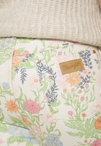 Vaqueros Wrangler con estampado floral sobre un fondo crema claro, con flores coloridas en tonos rosa, azul y verde, y una etiqueta de cuero marrón claro.