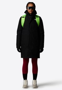 Svart parkas med høy krage, grønne stropper, røde leggins, svarte hansker og hvite joggesko. Har et strømlinjeformet design og reflekterende detaljer.