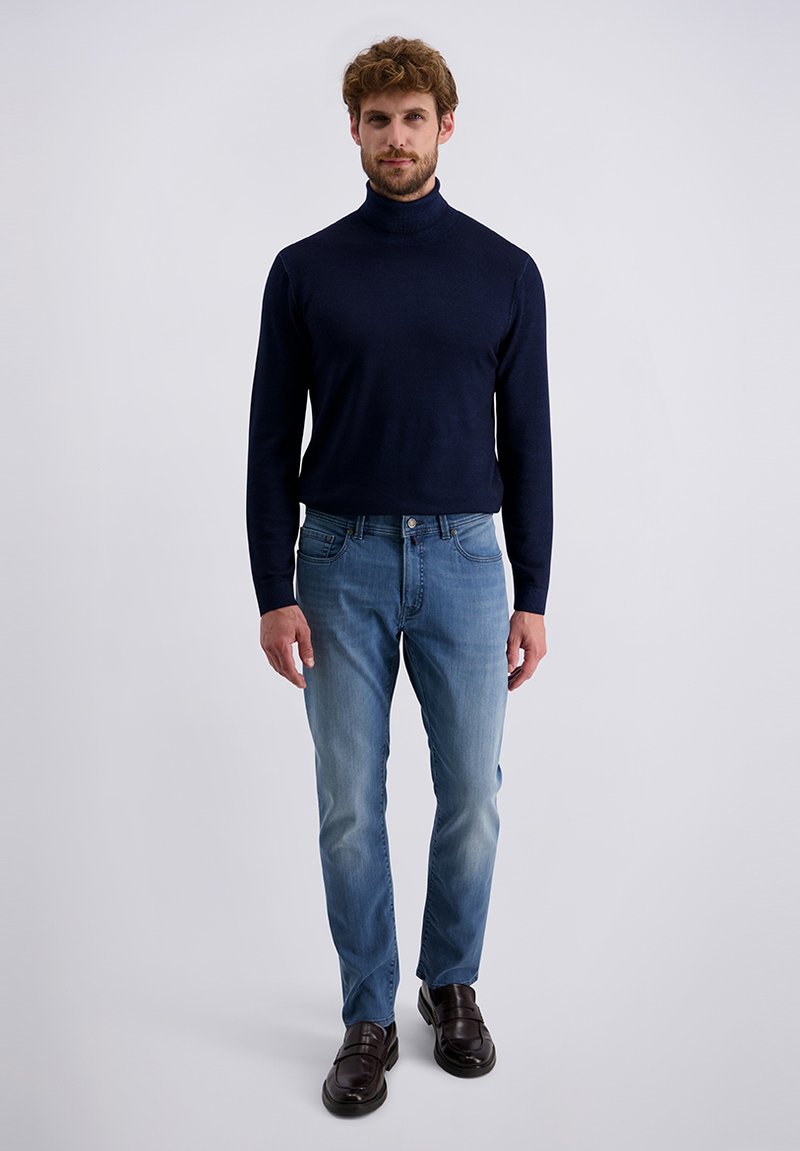 Mann steht in einem marineblauen Rollkragenpullover, hellblauen Jeans und dunkelbraunen Lederloafers vor einem hellgrauen Hintergrund.