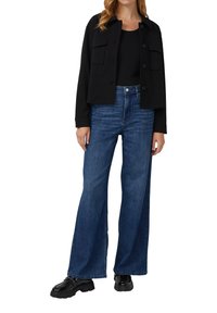 Zwarte cropped jas met twee borstzakken, gecombineerd met hooggezette, wijde, blauwe jeans. Schoeisel heeft glanzende zwarte zolen.