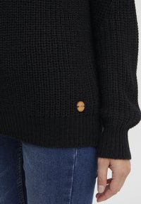 Schwarzer gestrickter Pullover mit einem strukturierten Muster, ausgestattet mit gerippten Bündchen und einem Holzlogo-Akzent am Saum. Kombiniert mit blauen Jeans.