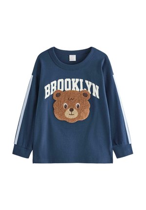 Lille barn med blondt hår iført en navyblå skjorte med en bjørneansigt og teksten "Brooklyn", løse lyseblå jeans og hvide sneakers.