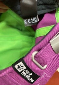 Paarse rugzakriem met metalen gesp, Jack Wolfskin-logo patch en een gedeeltelijk zichtbare zwart-witte CE UKCA-label aan de binnenkant van de groene voering.