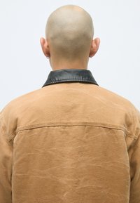 Brun kanvasjakke med krave i svart lær. Stoffet har en teksturert finish med rynkedetaljer, sett fra baksiden.