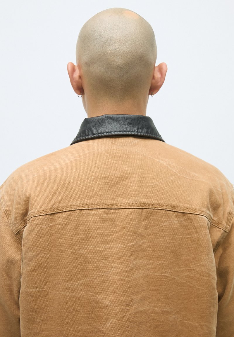 Braune Canvas-Jacke mit einem schwarzen Lederkragen. Der Stoff hat eine strukturierte Oberfläche mit gefälteltem Detail, gesehen von hinten.