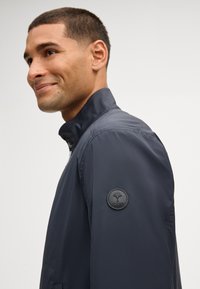 Mężczyzna w granatowej kurtce z podniesionym kołnierzem, lekko uśmiechnięty, z naszywką z logo na górnej części rękawa, zwrócony w lewo na jednolitym tle.