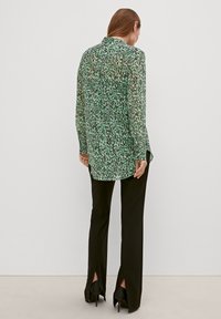 Blusa verde trasparente con un motivo sparso, colletto alto e maniche lunghe, abbinata a pantaloni neri sartoriali con spacco all'orlo.