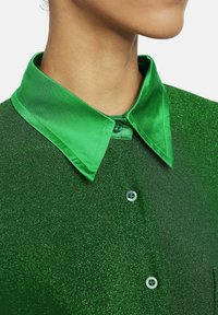 Grüne Bluse mit einem Satin-Kragen, aus strukturiertem Stoff und mit Knopfleiste. Die Farbe ist ein lebhaftes Grün mit einem subtilen Glanz.