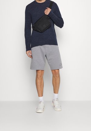 Granatowa koszula z długim rękawem, szare szorty z logo, czarna torba na ramię, białe trampki, białe skarpetki. Swobodny strój w luźnym kroju.