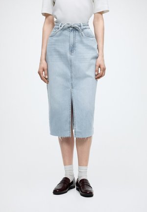 Frau trägt einen hellblauen Jeans-Midirock mit mittigem Schlitz, weißes kurzärmliges Oberteil, weiße Socken und dunkelbraune Loafers.