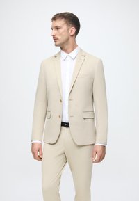 Šviesiai rudas kostiumas su pritaikytu švarku, turinčiu iškirptę, vieną krūtinės kišenę ir dvi mygtukas. Suderintas su baltu marškiniu ir diržu.