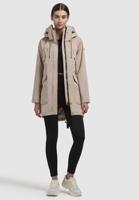 Giacca lunga beige con cappuccio, cerniera anteriore e coulisse; presenta due tasche laterali e un tessuto liscio e resistente all'acqua.