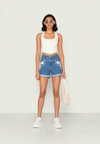 Μπλε τζιν σορτσάκια με άσπρη λουλουδάτη κεντημένη διακόσμηση, ψηλόμεσα και με ραμμένο στρίφωμα, συνδυασμένα με ένα εφαρμοστό λευκό crop top και παστέλ παπούτσια. Τα αξεσουάρ περιλαμβάνουν γυαλιά ηλίου και μια τσάντα tote.