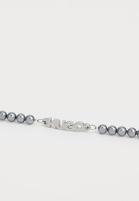 Grå pärlhalsband med metallisk HUGO-logotyp i mitten, växlande blanka och matta pärlor, säkrad med en silverkedja.