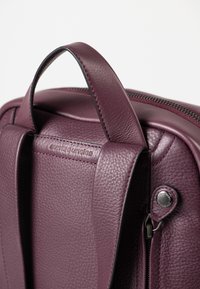 Paarse leren tas met een gestructureerde afwerking, voorzien van een handvat aan de bovenkant, contrasterende stiksels en een ritssluiting met metalen hardware.