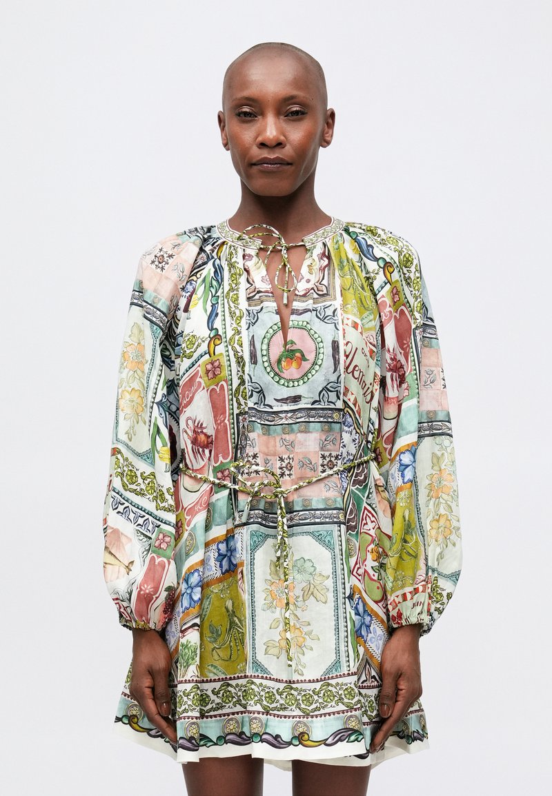 Persoană îmbrăcată într-o rochie colorată cu patchwork, având modele florale și fructe, mâneci lungi și puffate, precum și o funda la gât și la talie.