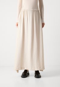 Cream-colored maxi skirt made from lightweight fabric with a textured finish. Features an elastic waistband and falls to ankle length. Black shoes below.