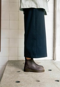 Botas de tobillo marrón oscuro con un acabado de cuero liso, paneles laterales elásticos y una suela resistente, combinadas con una falda larga negra.
