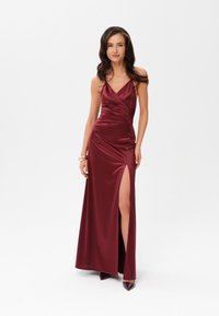 Burgundská saténová večerní šaty s hlubokým výstřihem do V, nařasenými detaily a bočním rozparkem. Má tenké ramínka a přiléhavý střih, doplněné o špičaté podpatky.