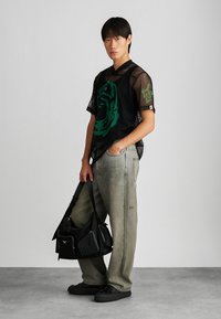 Juodas permatomas per didelis marškinėliai su žalia grafika, laisvi pilki džinsai su nedideliais dėmėmis, ir juoda kuprinė su keliomis kišenėmis bei užtrauktukais.