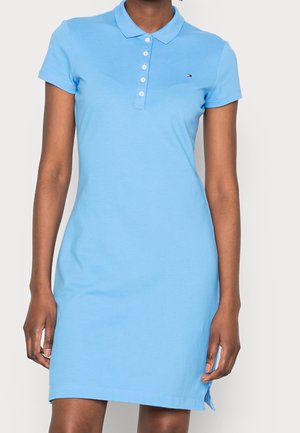 Woman wearing a light blue, short-sleeve polo dress with white buttons and a small logo on the left chest.