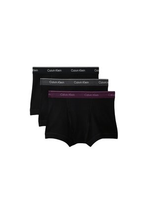 Gespiereerde man staat op blote voeten met gekruiste armen, draagt zwarte Calvin Klein boxershorts tegen een effen lichtgrijze achtergrond.