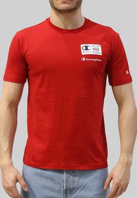 T-shirt rosso a maniche corte realizzato in cotone con scollo rotondo. Presenta un logo 'Champion' sul petto e un piccolo logo sulla manica.