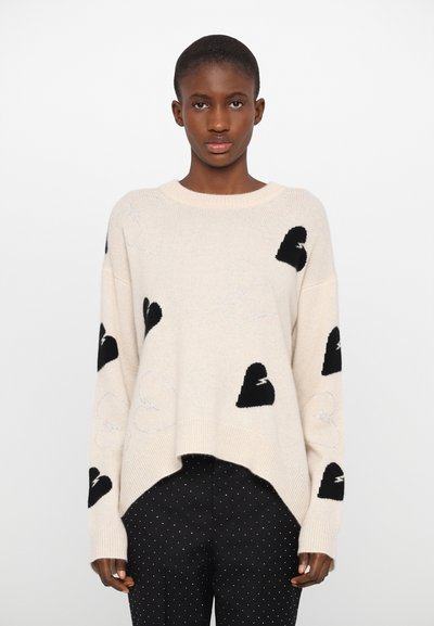 Pull en tricot couleur crème avec des motifs de cœurs noirs et des accents de fil argenté. Coupe loose, col rond et ourlet côtelé.