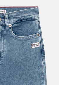 Světle modré denimové džíny s přední kapsou, kovovým knoflíkem na zapínání a páskou s nápisem "HILFIGER 1985" na boku. Červené prošívání jako akcent.