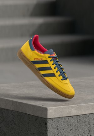 Κίτρινο αθλητικό παπούτσι Adidas Spezial με μπλε ρίγες, μπλε κορδόνια, κόκκινη εσωτερική επένδυση και καφέ σόλα, που βρίσκεται πάνω σε τσιμεντένια επιφάνεια με σκάλες στο φόντο.