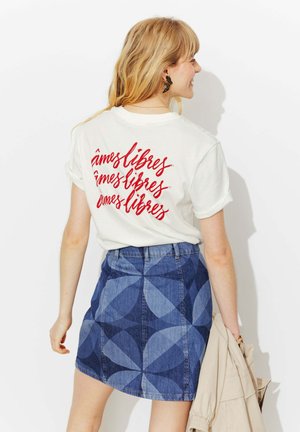 Femme blonde vue de dos portant un t-shirt blanc avec le texte rouge « âmes libres » et une jupe en denim à motifs géométriques bleus, tenant une veste beige.