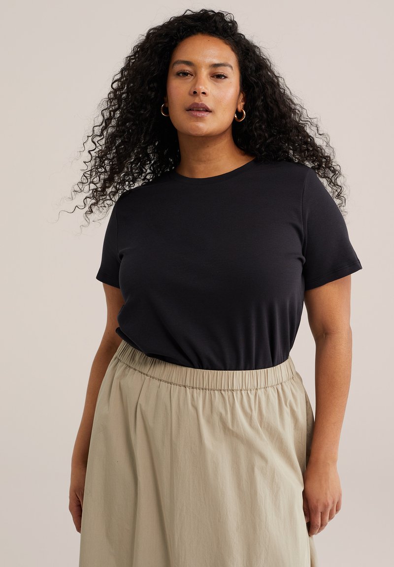 Žena s kovrčavom kosom koja nosi zlatne naušnice u obliku prstena, crni kratki top i bež suknju visokog struka na jednobojnoj pozadini.