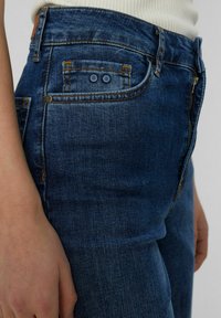 Mörkblå denimjeans med en figurnära design, som har en framficka, bakfickor med detaljer och orangea sömmar.