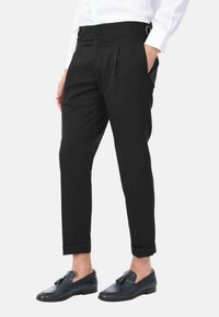 Pantaloni neri su misura con pieghe, orli risvoltati e una vita ampia con bottoni decorativi e un regolatore laterale.