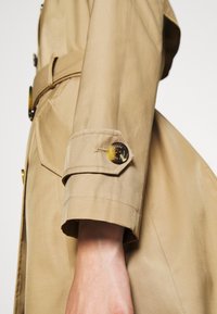 Beżowy płaszcz trencz z paskiem, z detalem z guzika na rękawie i gładką teksturą. Tkanina ma delikatny połysk.