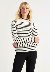 Stribet sweater i creme og sort, med ribbet tekstur, høj hals og gylden broderi på brystet. Sammen med sorte leggings.