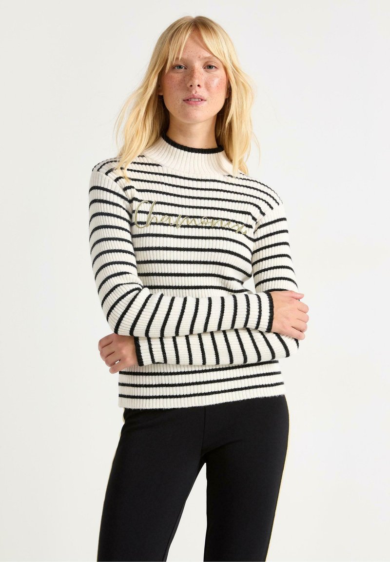 Stribet sweater i creme og sort, med ribbet tekstur, høj hals og gylden broderi på brystet. Sammen med sorte leggings.